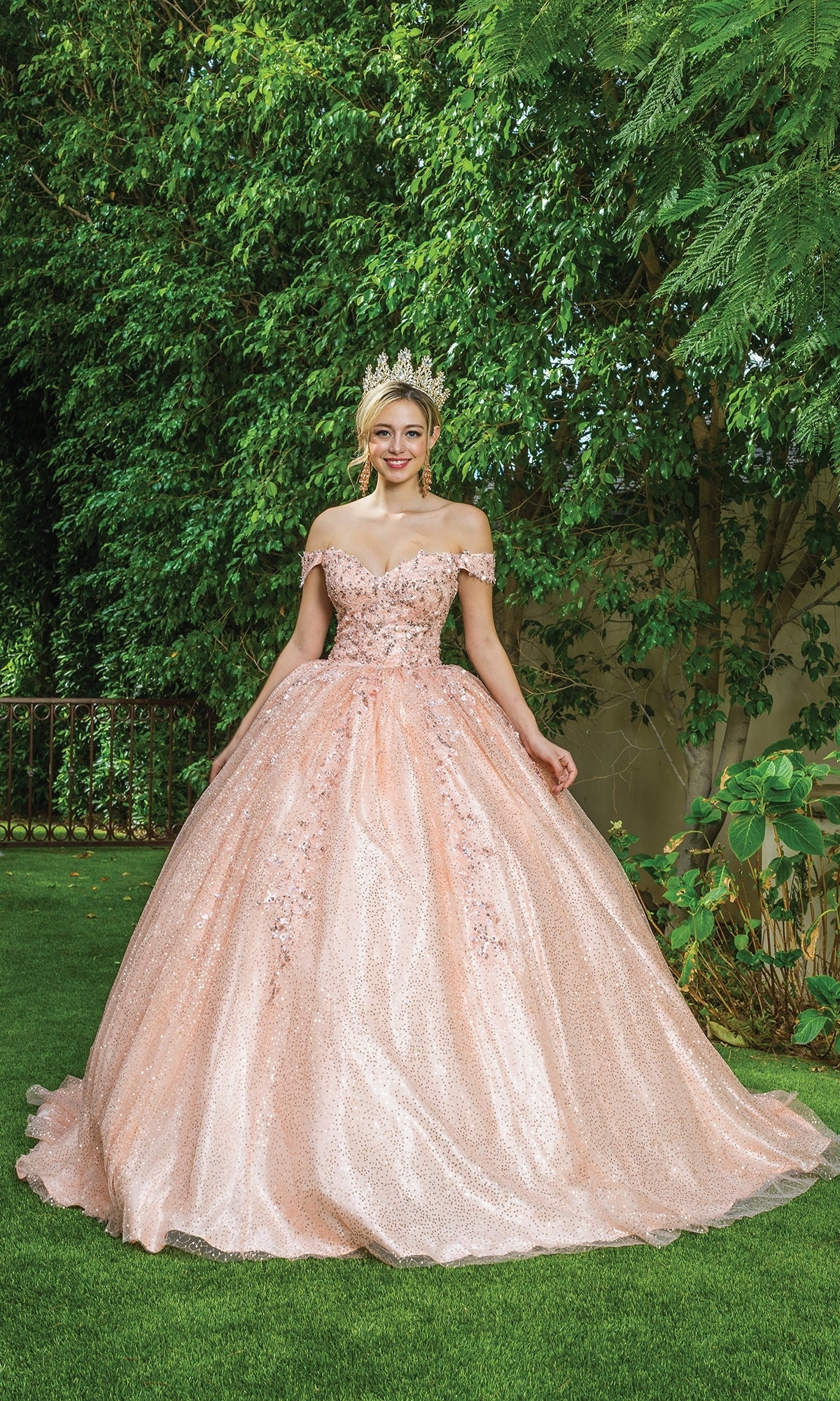 A young woman smiles at the camera, standing on grass in front of green foliage, wearing the Quinceanera Dress 1595 by Dancing Queen—a pink off-the-shoulder ball gown with a sweetheart neckline and a tiara.